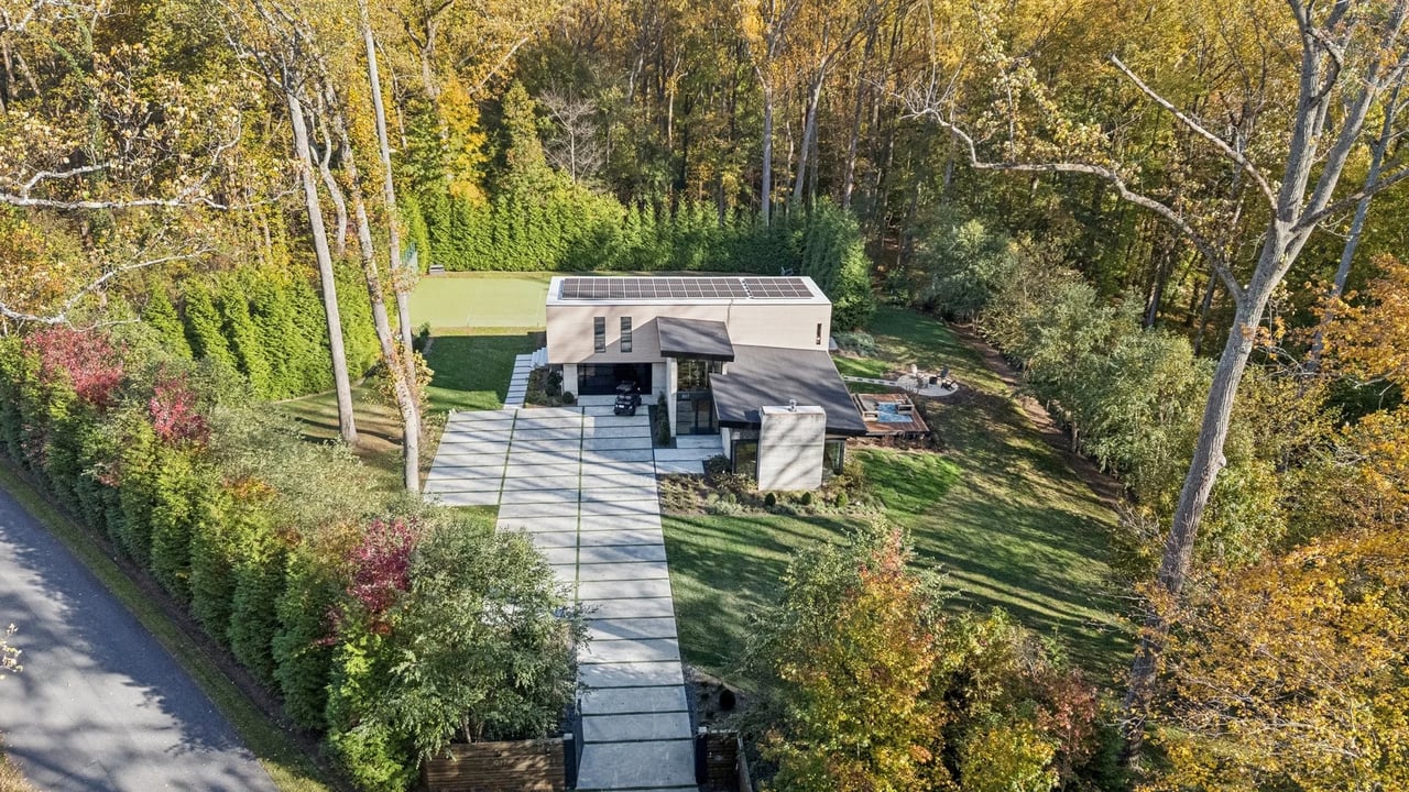 Aerial view of a modern home with rooftop solar panels and a geometric driveway among autumn trees.