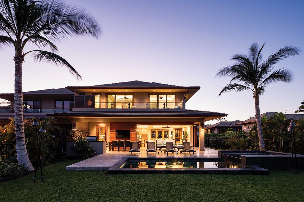 Modern two-story home with an illuminated patio, swimming pool, and palm trees at dusk.