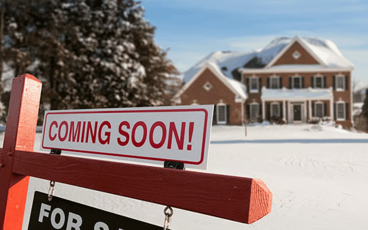 Snow-covered brick house behind a red “Coming Soon!” real estate sign.