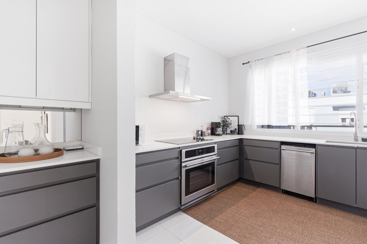 Modern kitchen with gray cabinets, stainless-steel appliances, and a large curtained window.