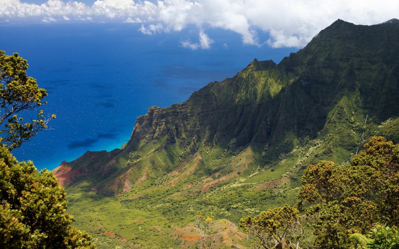 Rugged green coastal mountains above deep blue ocean beneath a cloudy sky.
