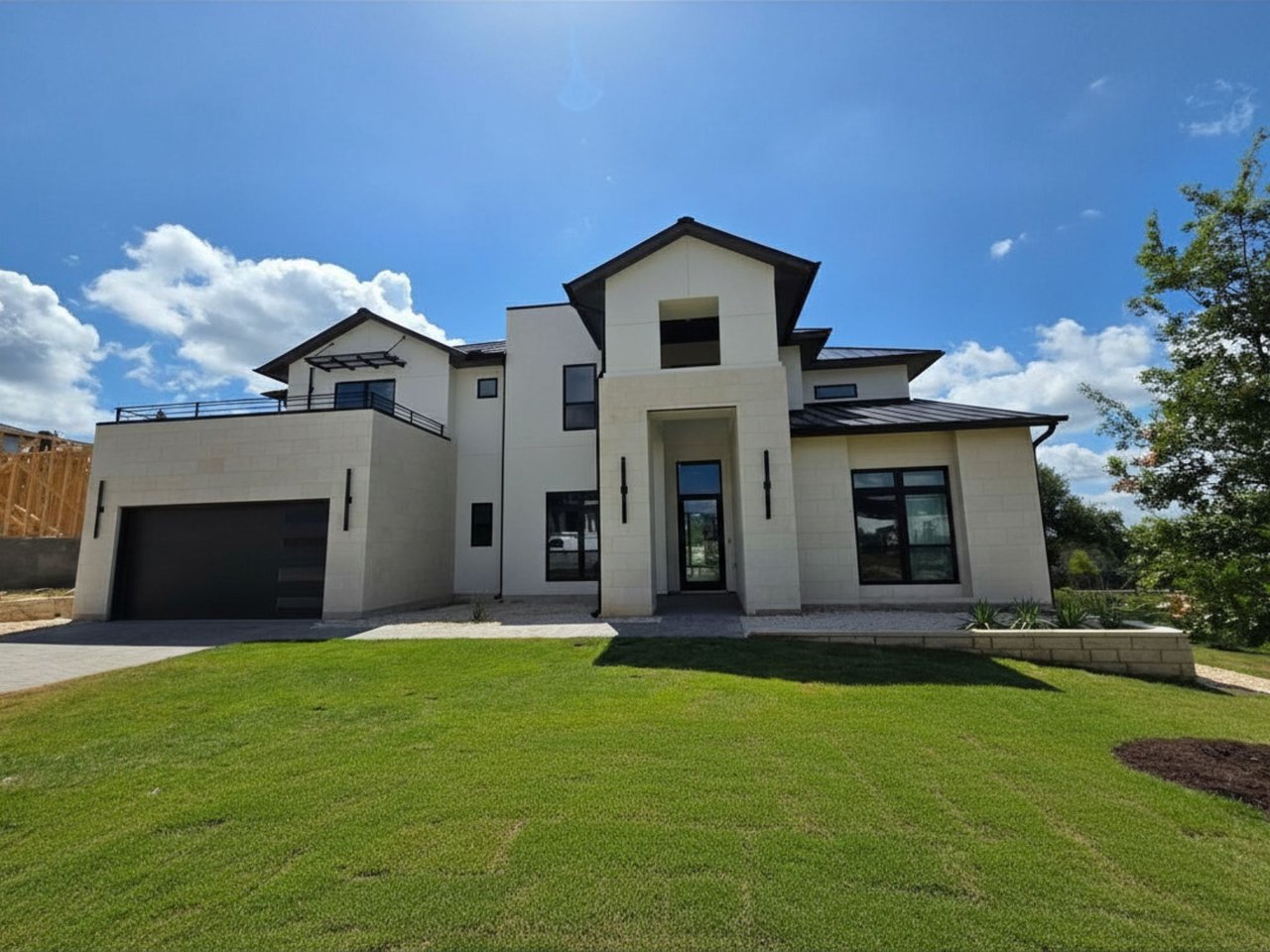 Modern two-story home with pale stone facade, black garage door, upper balcony, and green lawn.
