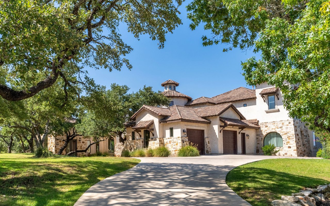 Large stucco and stone house with terracotta tile roofs behind a curved driveway and mature trees.