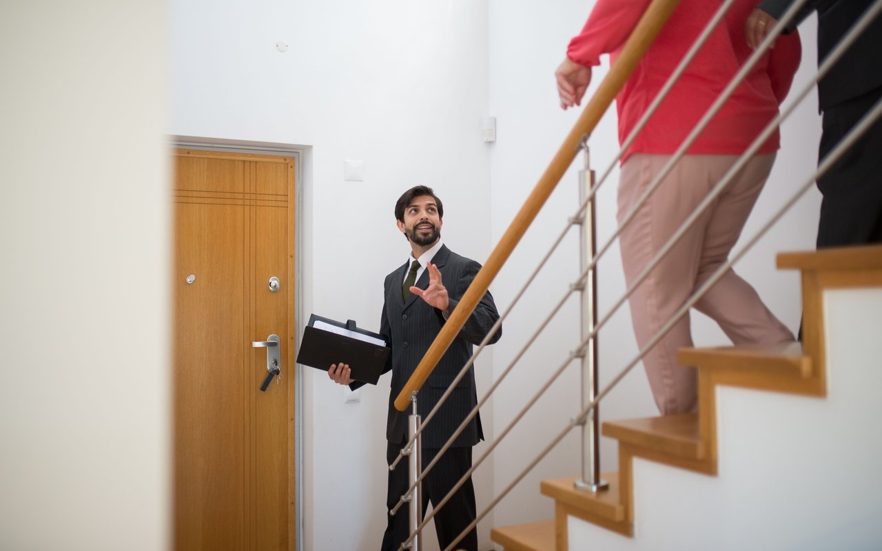 A real estate agent with a folder speaks to a client on a wooden staircase.