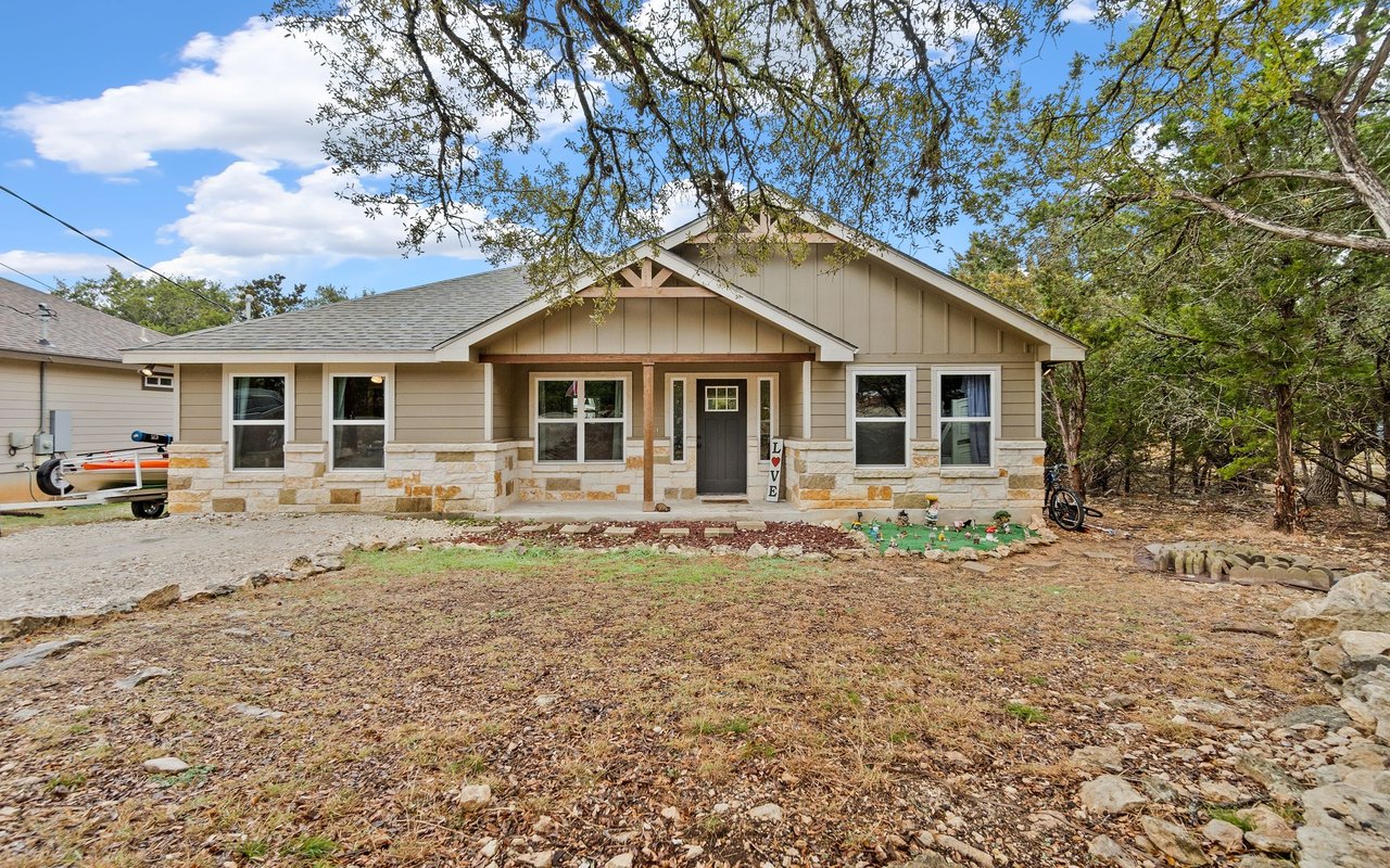 Single-story beige home with a stone facade, covered porch, and wooded yard.