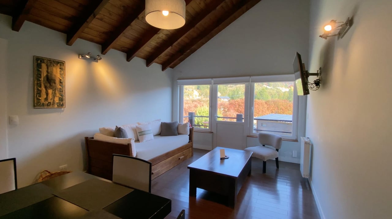 Living room with a white sofa, exposed timber ceiling, and large windows overlooking autumn-colored trees.