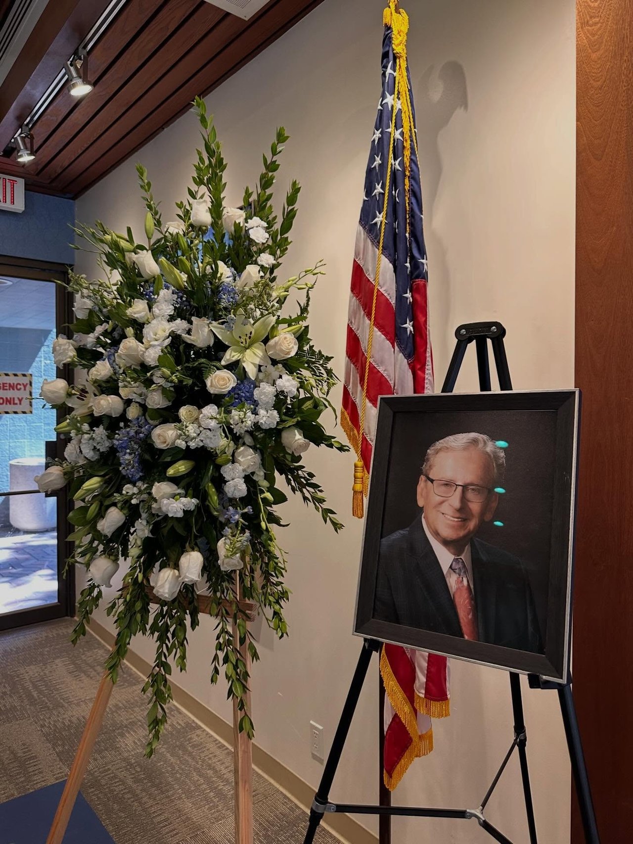 Framed portrait of Al Shkoler beside a large floral arrangement and American flag.