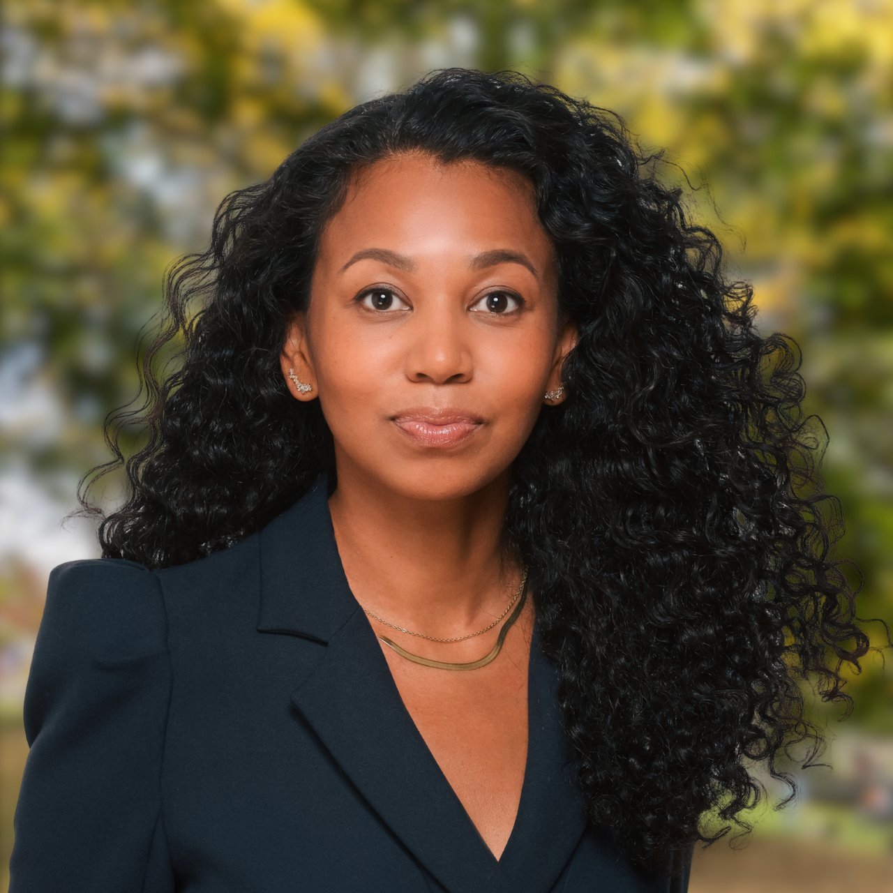 Nadirah Chestnut in a dark blazer, with long curly hair against a blurred leafy background.