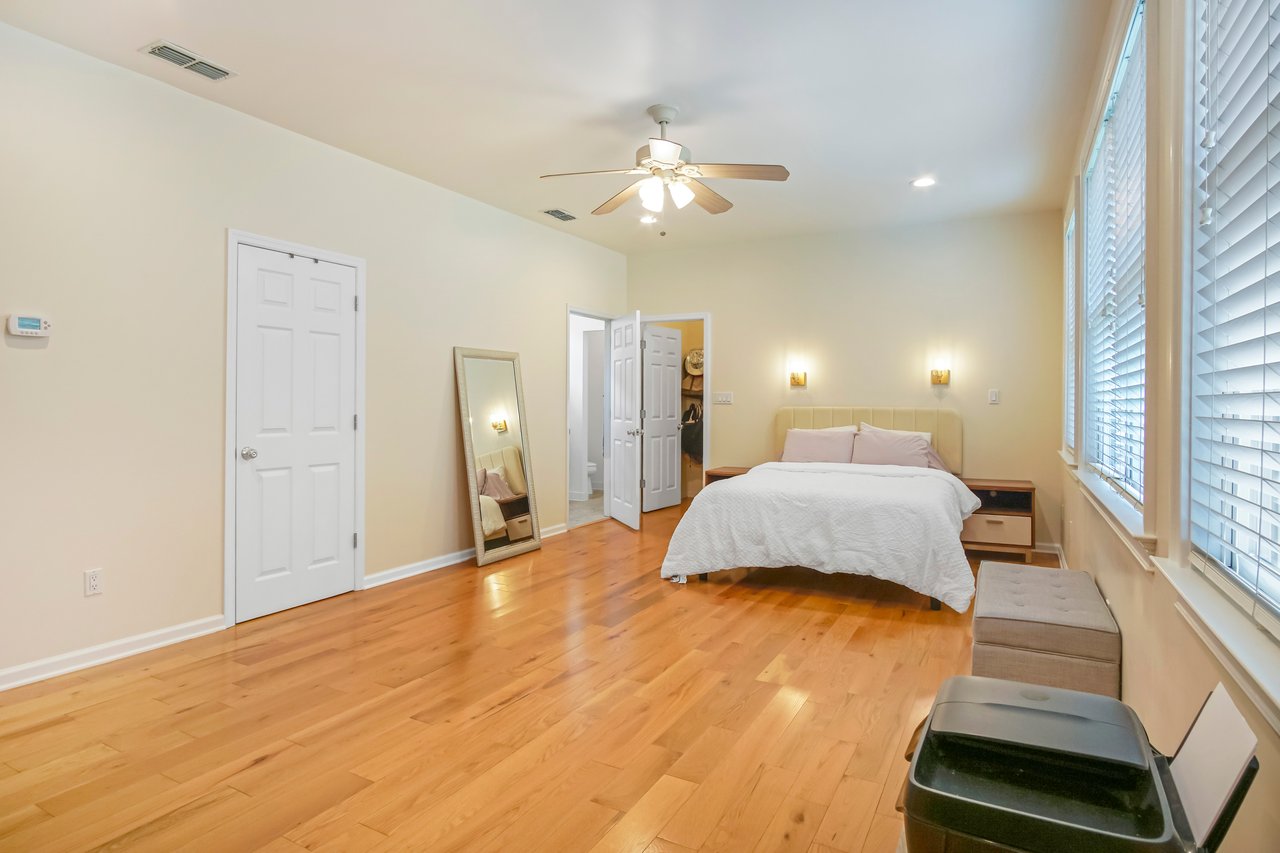 Spacious bedroom with wooden floor, cream walls, and a ceiling fan. Features a bed, wall mirror, and window blinds, evoking a calm and airy ambiance.