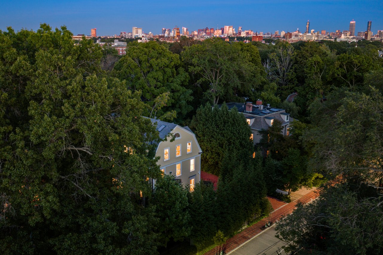 Victorian house among mature trees, with a brick walkway and city skyline at dusk.