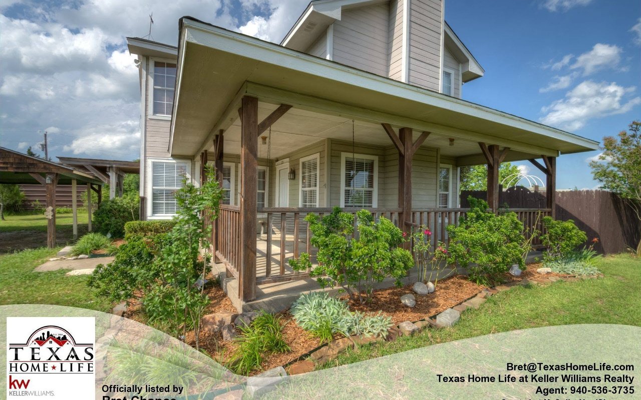 Two-story house with a broad covered porch, timber supports, landscaped garden beds, and a grassy yard.