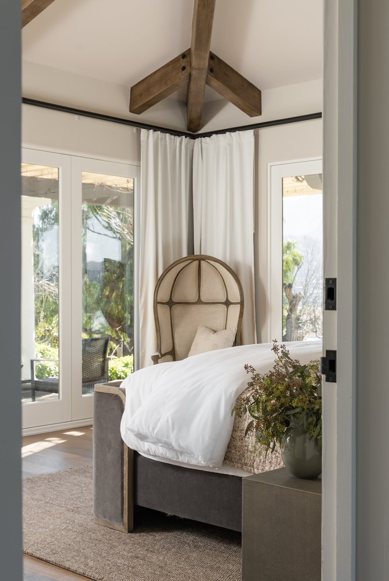 Staged bedroom with a gray upholstered bed, arched headboard, exposed wood beams, and glass doors.