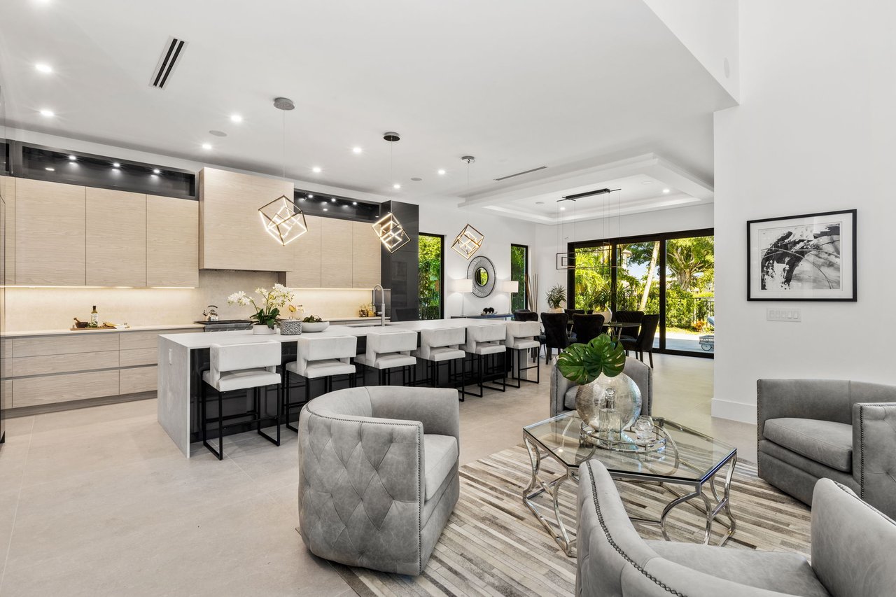 Open-plan kitchen and living area with a long island, gray seating, and windows overlooking tropical greenery.