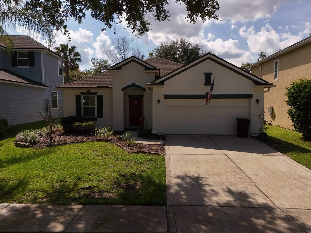Single-story beige house with a two-car garage, American flag, landscaped lawn, and concrete driveway.