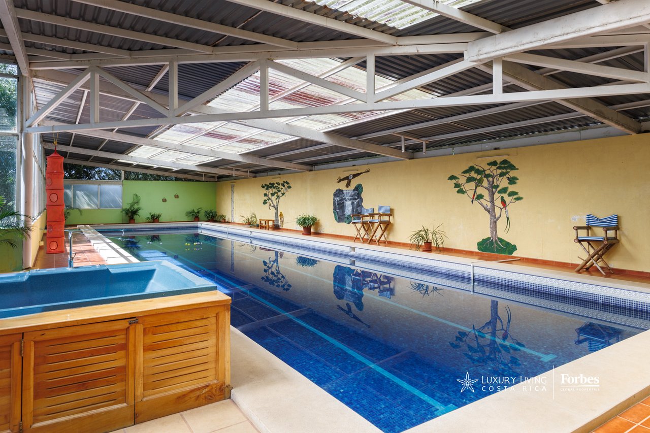 Covered indoor swimming pool with a small spa pool and director chairs along a mural-painted wall.