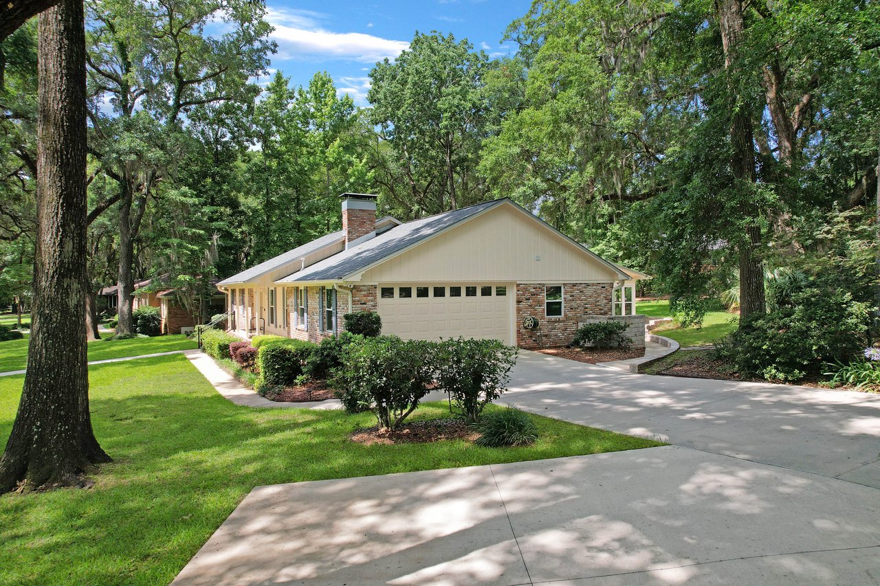 A charming one-story brick house with a garage surrounded by lush green trees and bushes. The driveway is wide, and the sky is clear and blue.