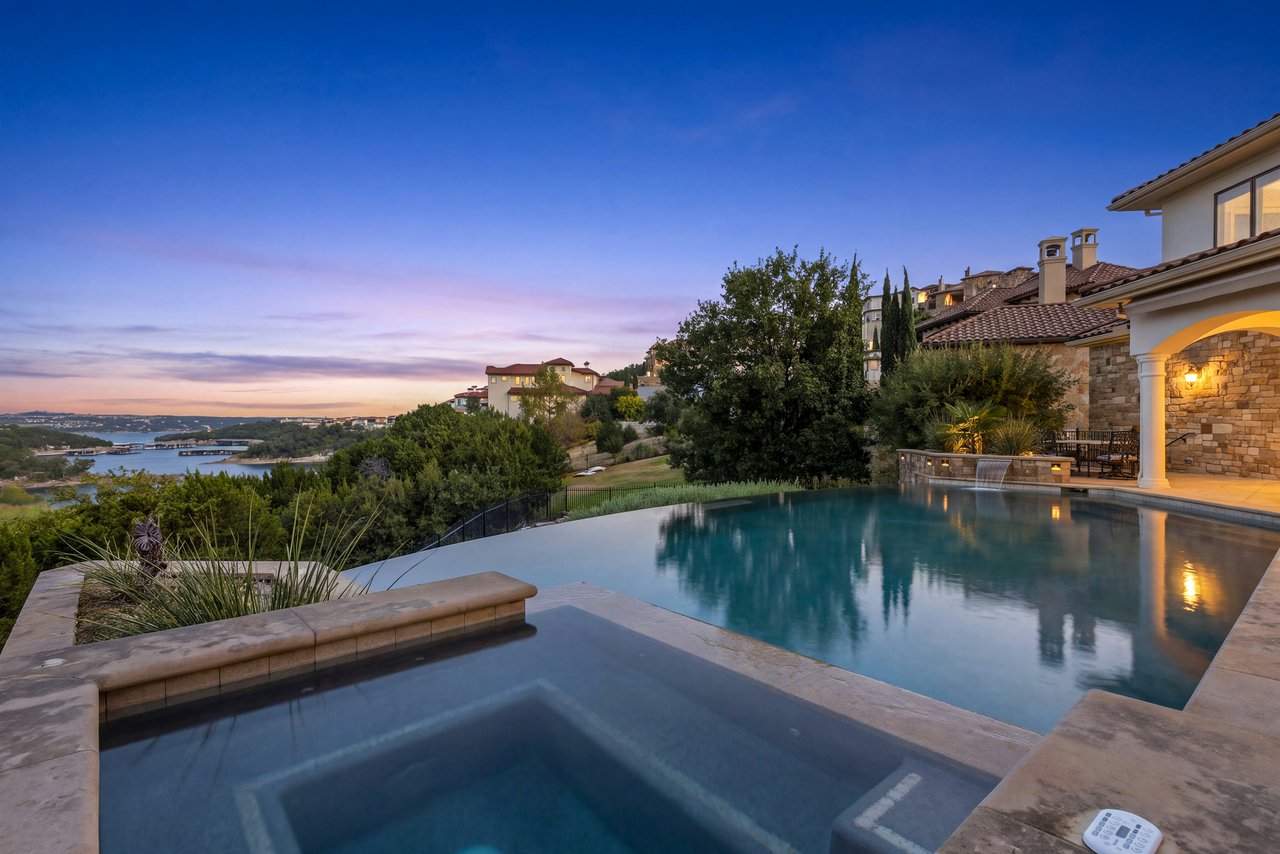 Home’s infinity pool and spa overlook Lake Austin at dusk, beside a warmly lit stone patio.