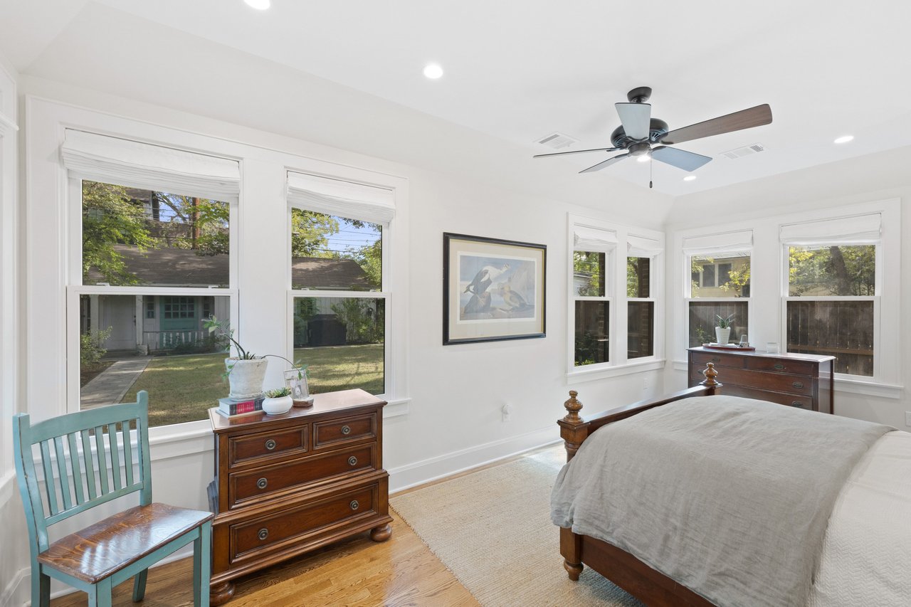 Bedroom with a large bed, hardwood floors, ceiling fan, and windows overlooking a yard.