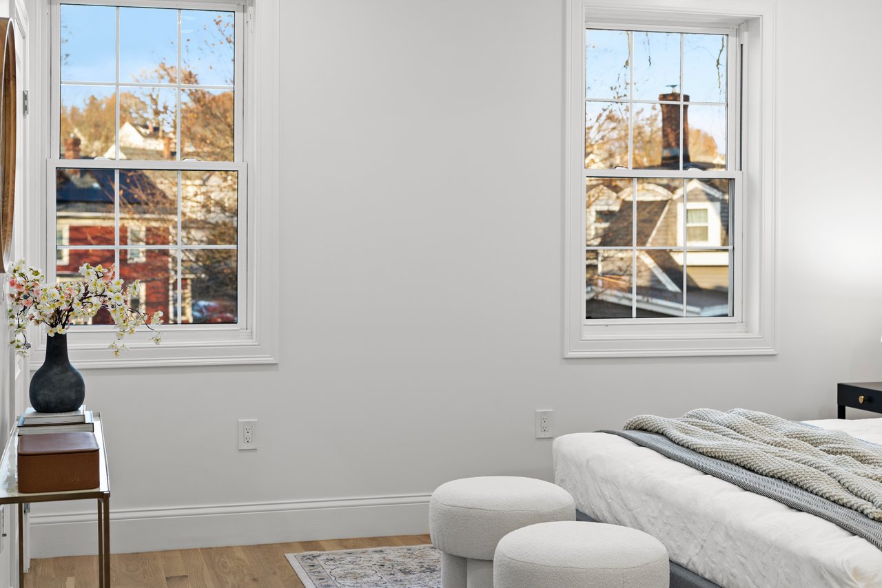 Staged bedroom with two large windows, a white bed, and upholstered ottomans.