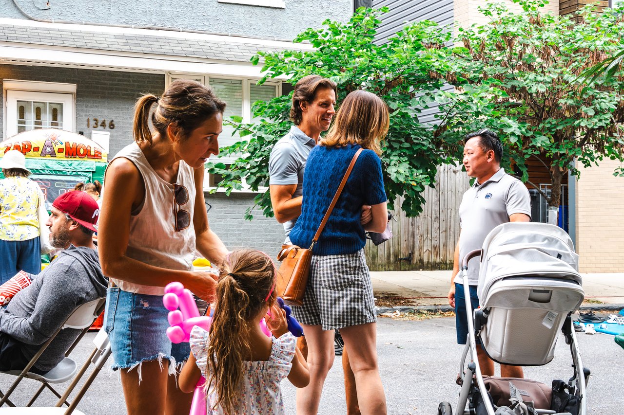 Neighbors mingle at an outdoor block party as a child holds a pink balloon sculpture.
