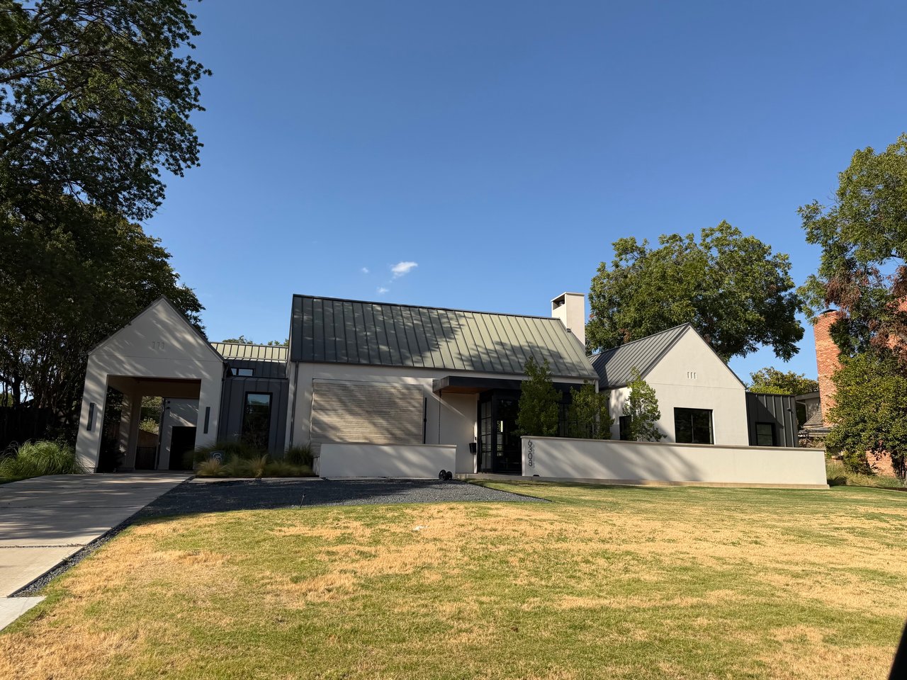 Modern white home with dark standing-seam roof, broad lawn, and mature shade trees.