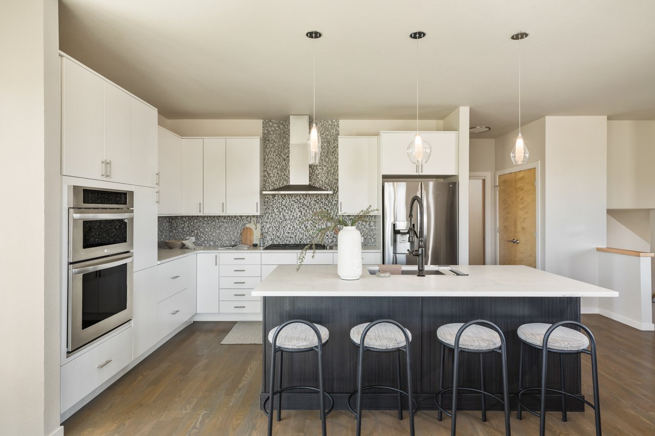 Kitchen with white cabinetry, a dark island with four stools, wall ovens, and a chimney hood