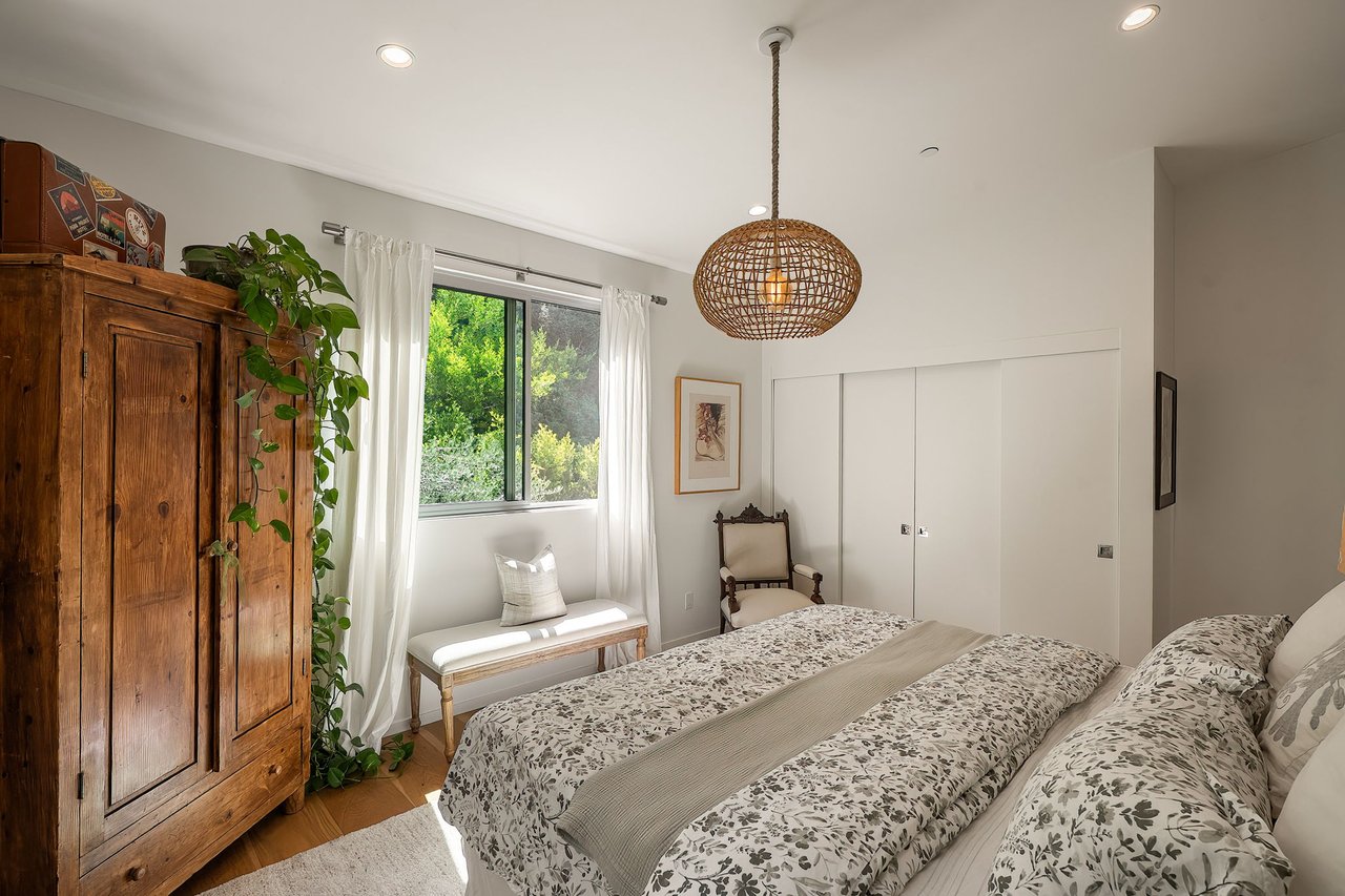 Bedroom with a floral bedspread, wooden armoire, and window overlooking greenery.
