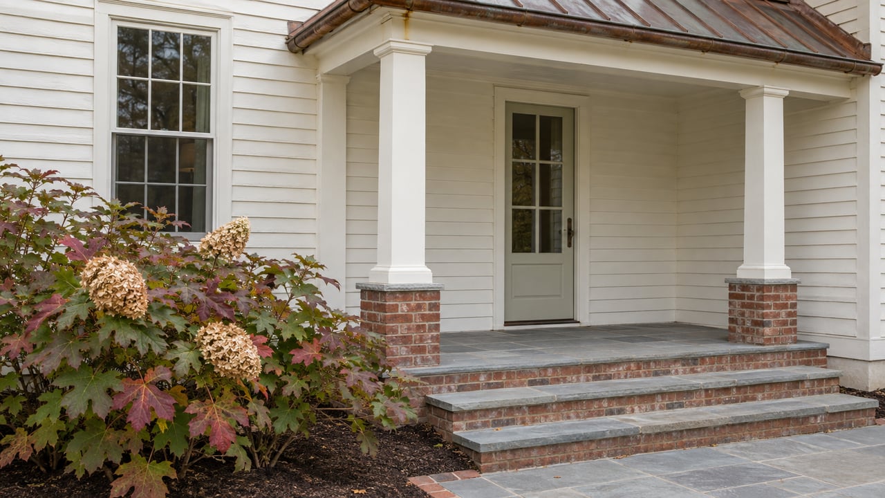 Traditional house porch with ivory clapboards, brick piers, bluestone steps, and burgundy-leaved oakleaf hydrangea.