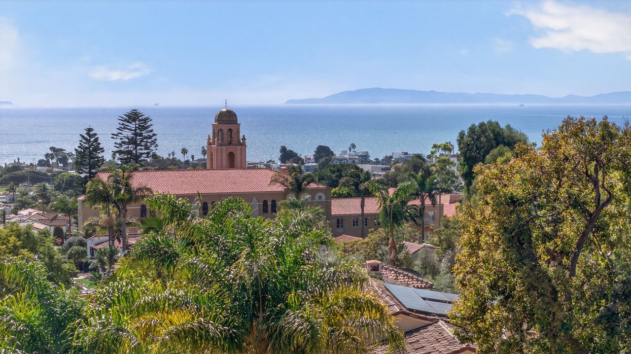 Coastal neighborhood with a bell tower and palm trees overlooking the ocean and distant islands.