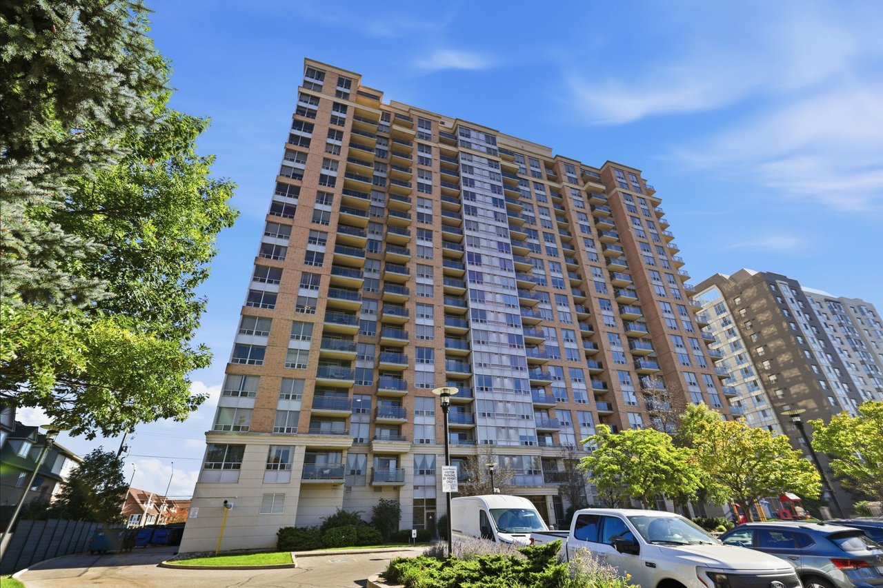 Brick condominium tower with rows of balconies, leafy trees, and parked vehicles.
