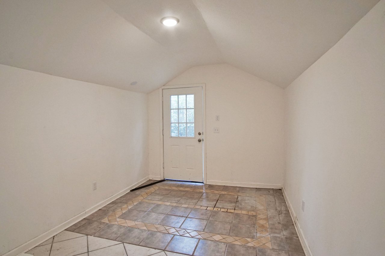Small room with slanted ceiling and white walls. Patterned tile flooring leads to a closed white door with a window, creating a minimalist, bright atmosphere.