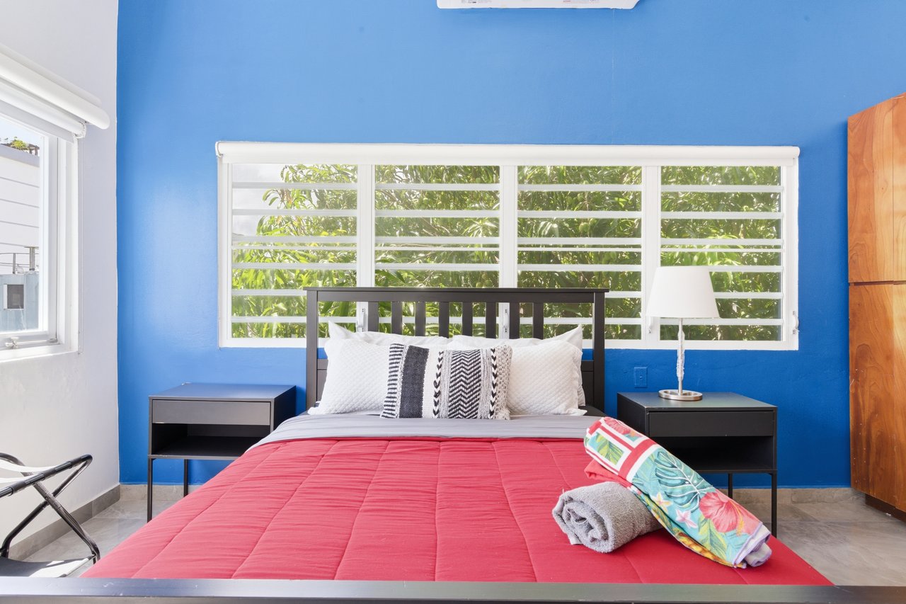 Bedroom with a blue accent wall, wide louvered window, and red quilted bed.