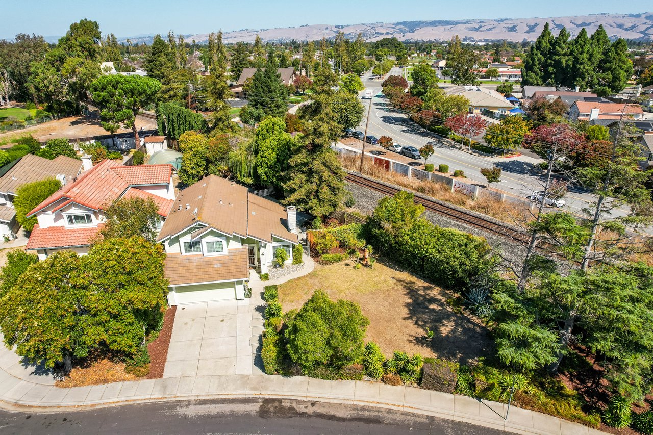 Aerial view of a single-story home with a tan roof, dry yard, and railroad tracks nearby.