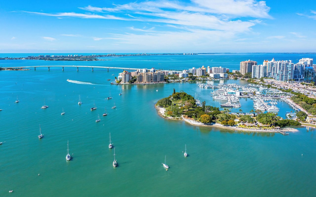 Aerial view of Sarasota Bay, with sailboats, a marina, downtown buildings, and a long bridge.