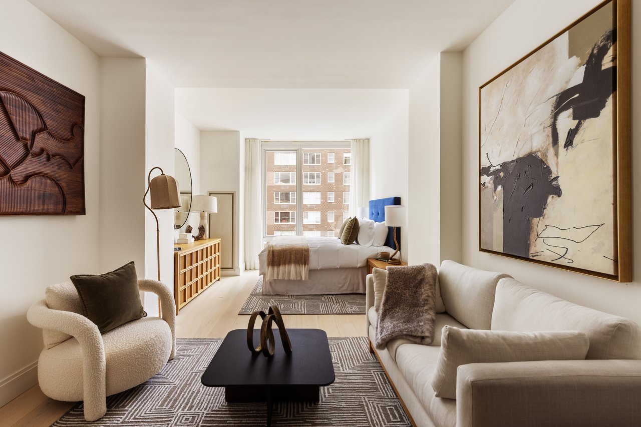 Staged studio apartment with a white sofa and armchair facing a bed beside windows overlooking a brick building.