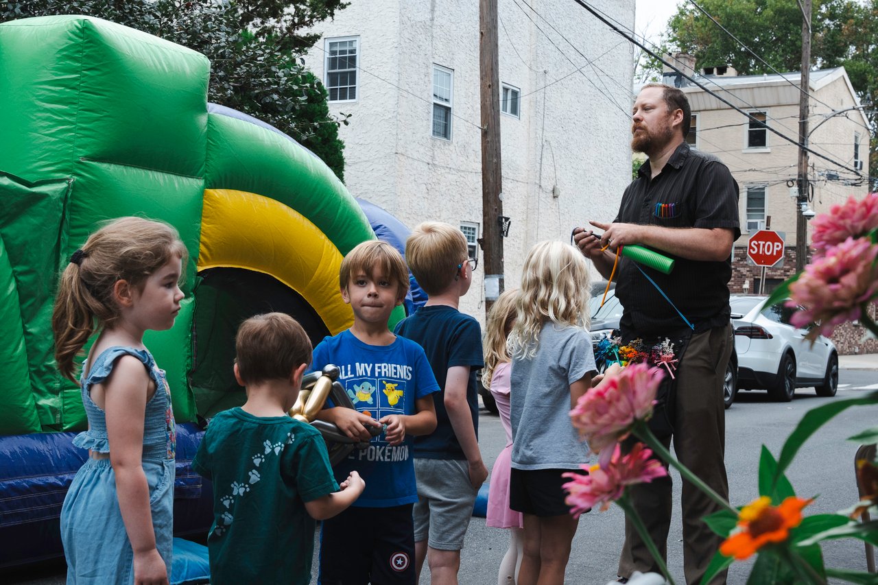 Children watch a man twist balloons beside a colorful bounce house at a neighborhood block party.