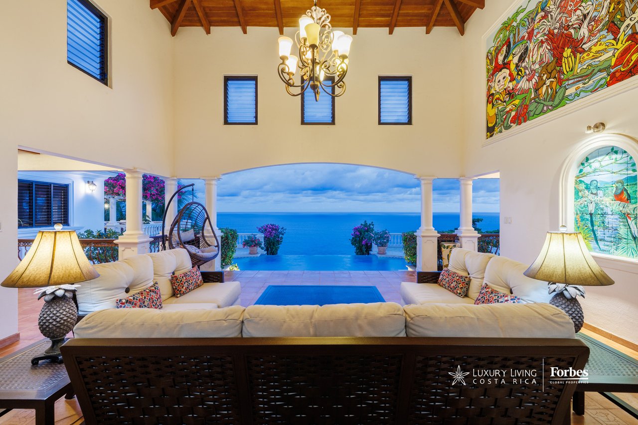 Living room opening onto an infinity pool and blue ocean view, with a vaulted timber ceiling.