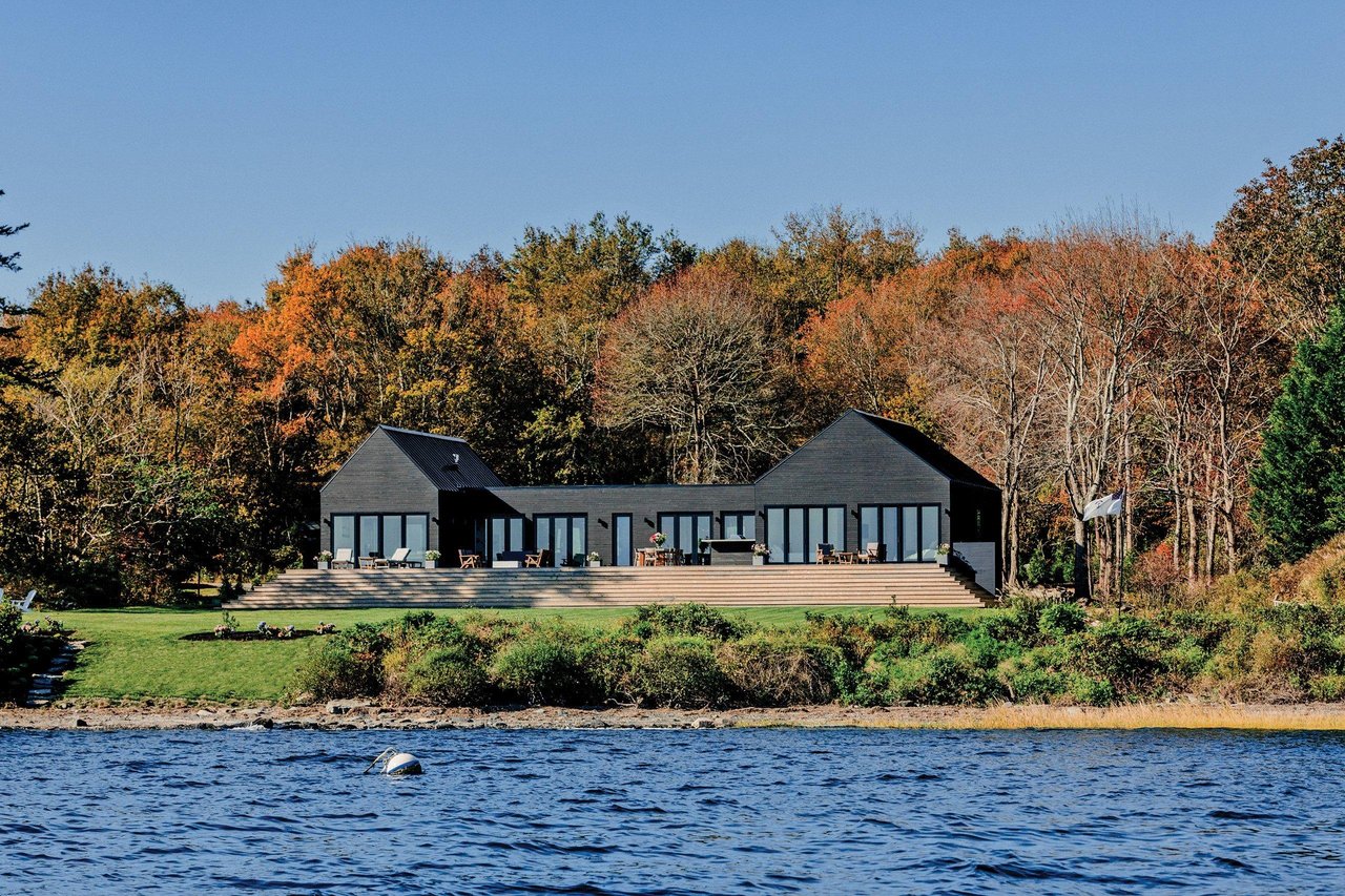 Modern lakeside home with a broad deck, autumn trees, and water in the foreground.