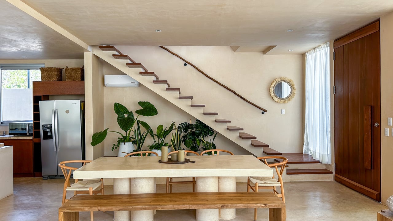 Dining area with stone table and wooden bench beneath an open staircase, beside a leafy indoor garden.