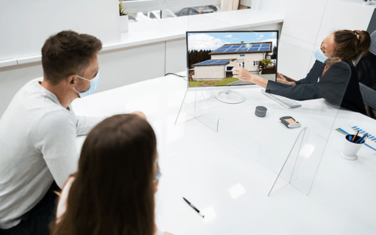 Masked clients view a solar-powered home on a monitor during a real estate consultation.