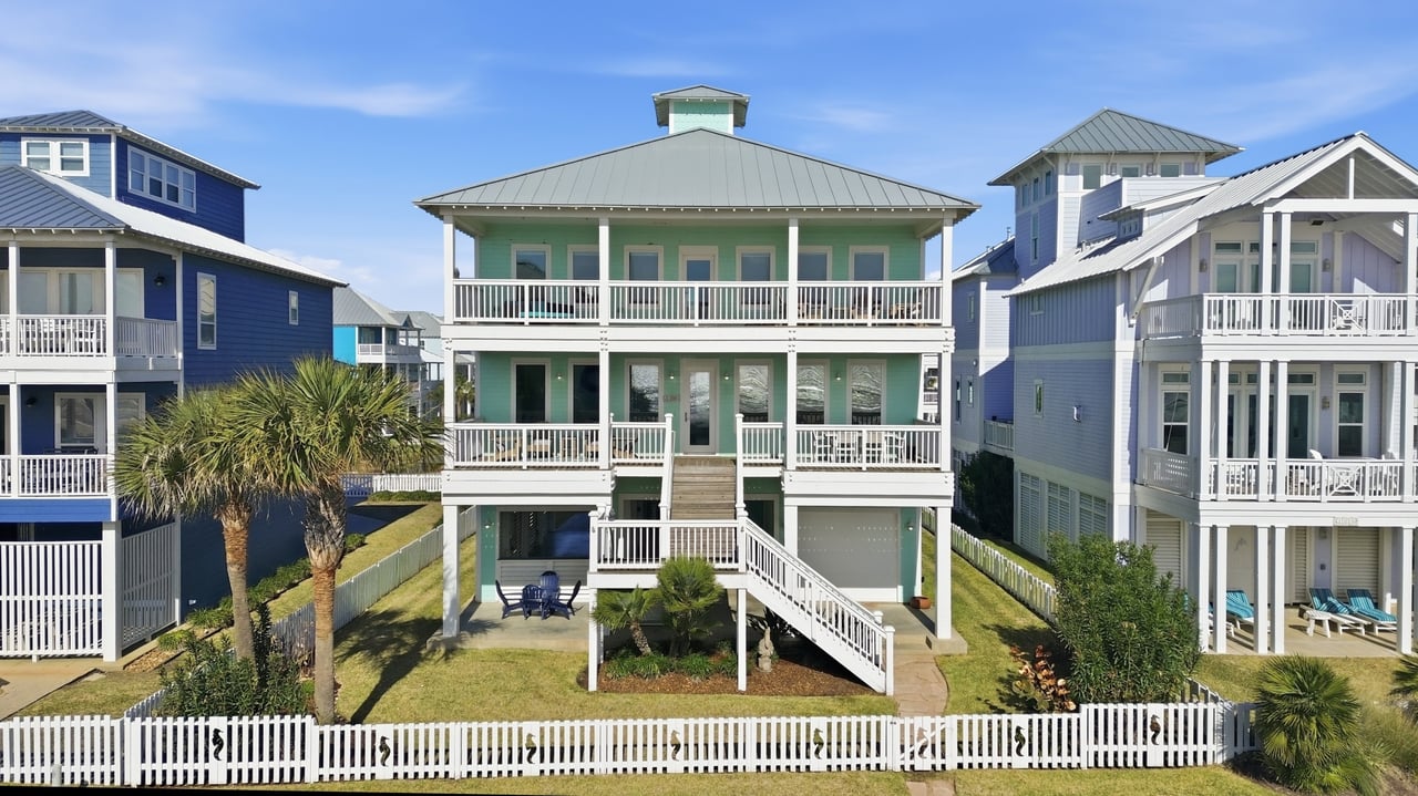 Mint-green beach house with broad white balconies, exterior stairs, and a white picket fence.