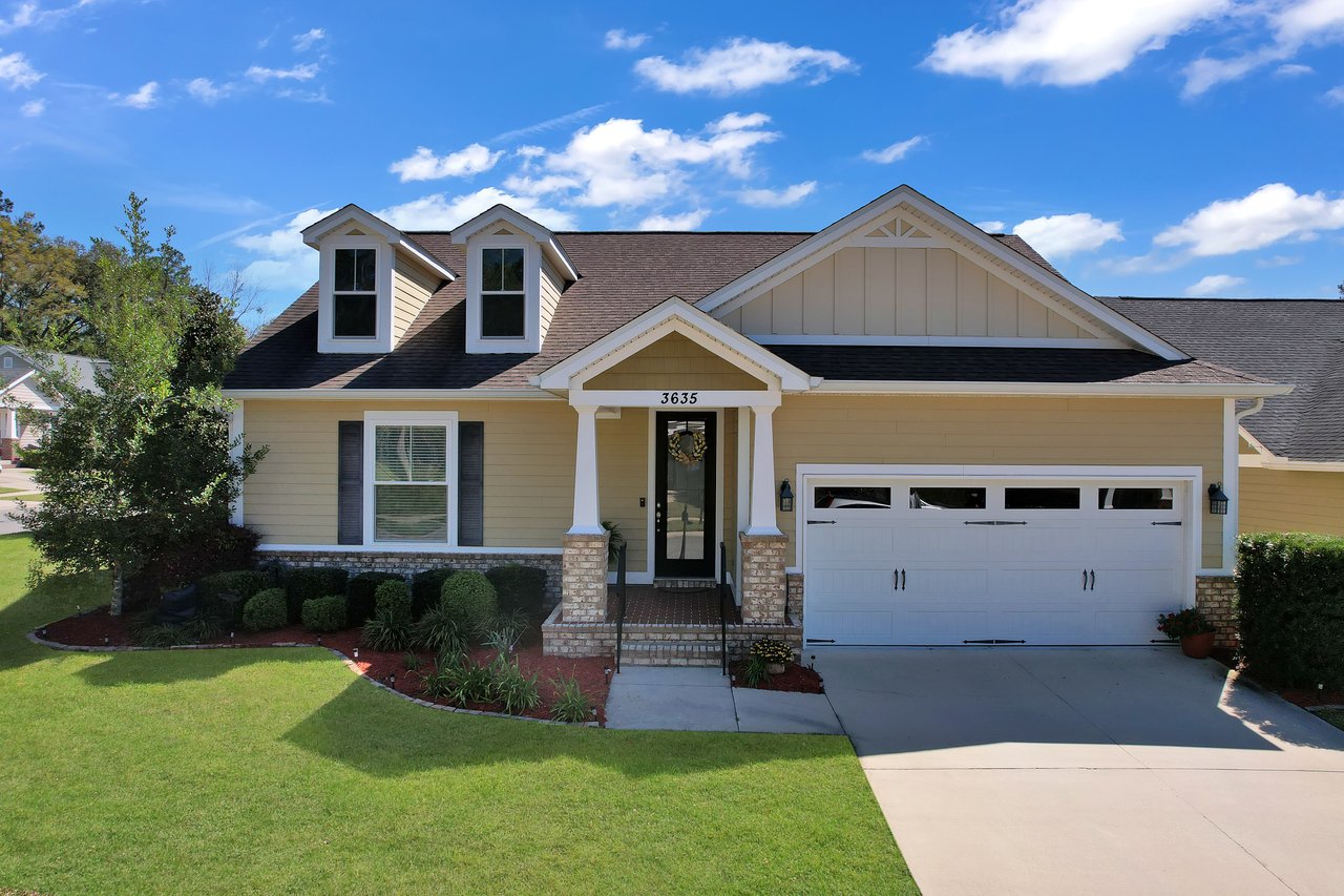 Single-story house with yellow siding, white trim, and a black roof. Features two dormer windows, a welcoming front porch, and a garage. Bright, sunny day.