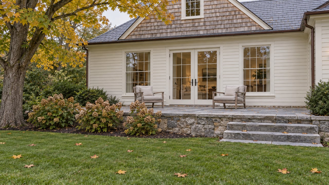 Autumn lawn leading to a limestone terrace and cream-clapboard home with cedar shingles, tall windows and silvered teak chairs.