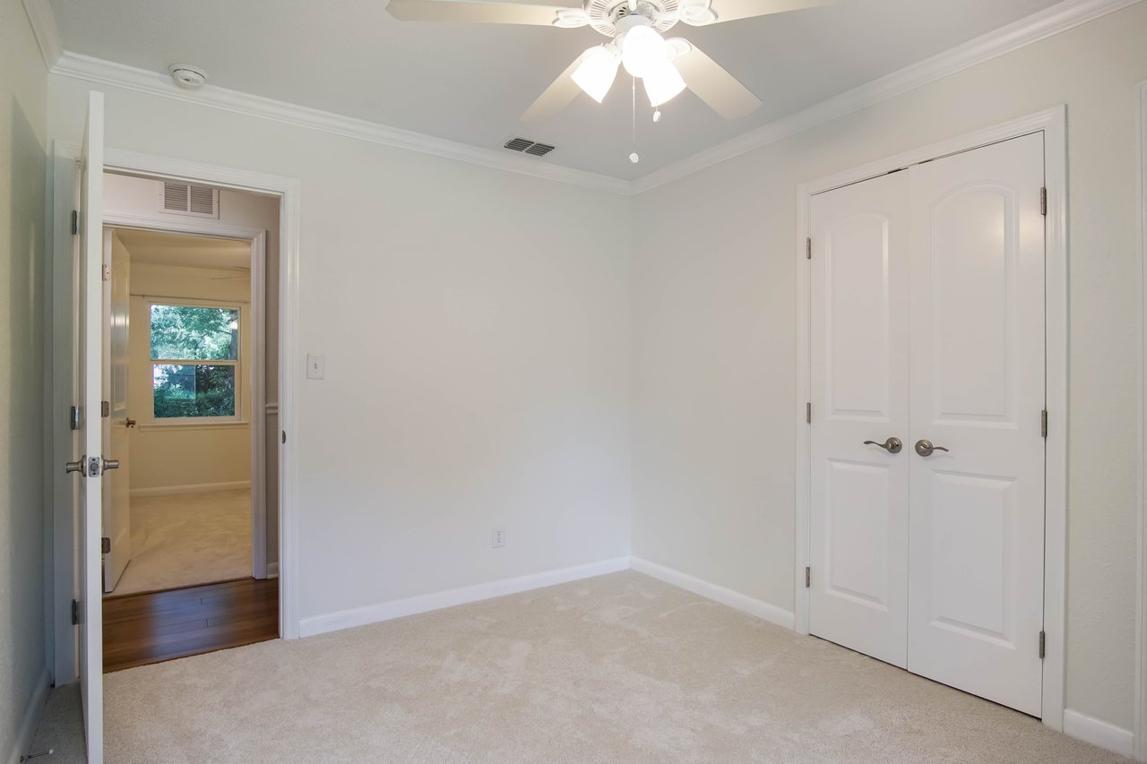Small, empty room with light beige walls and carpet. A ceiling fan with lights hangs above. A doorway opens to a room with hardwood flooring and a window. A closed double-door closet is on the right.
