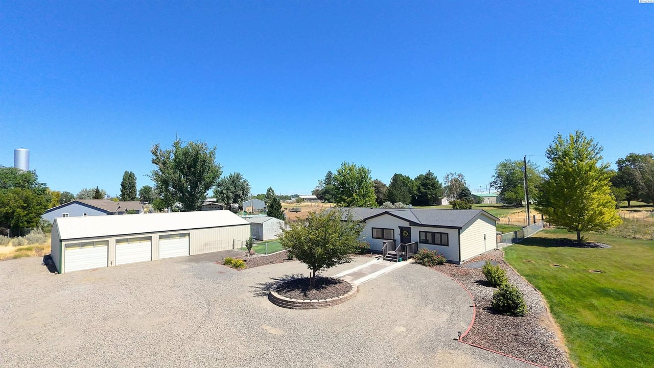 Single-story country home with a detached four-car garage, circular gravel driveway, mature trees, and expansive property in Burbank, Washington.