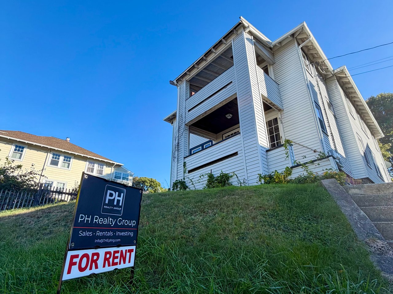 White multi-unit house on a grassy slope, with a PH Realty Group for-rent sign in front.