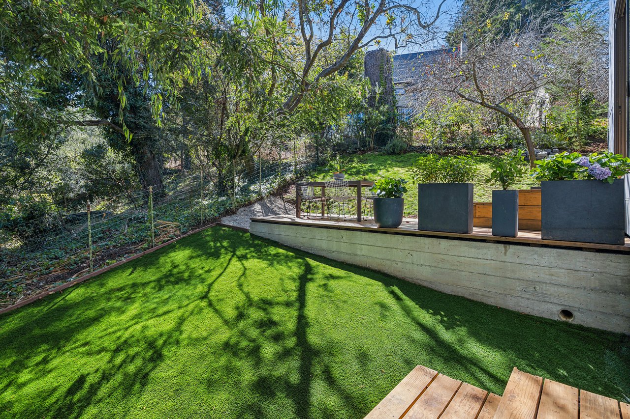 Landscaped backyard with artificial turf, mature trees, and a raised patio with outdoor dining furniture.