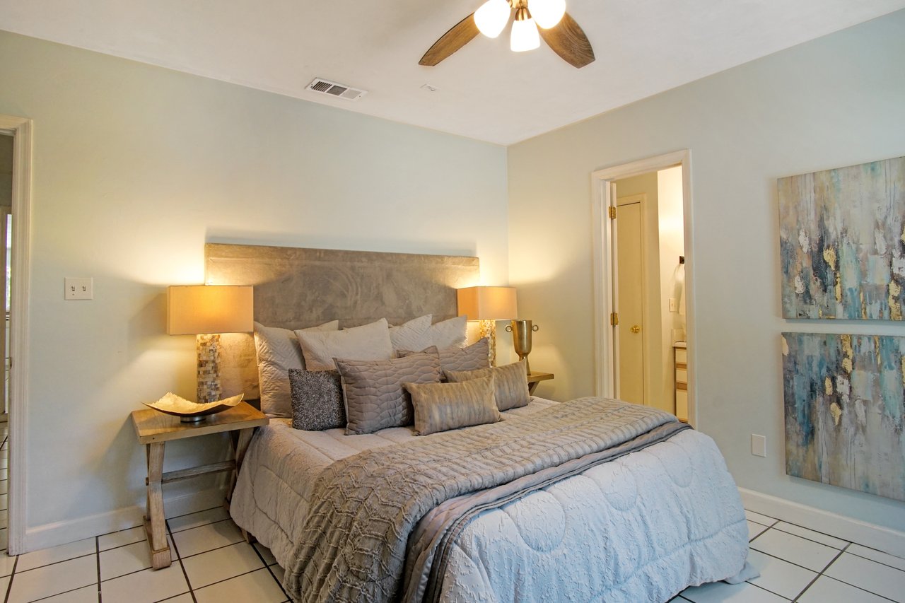 Cozy bedroom with a grey bed, layered pillows, and matching quilt. Two bedside lamps emit warm light. Abstract art decorates the wall; serene ambiance.