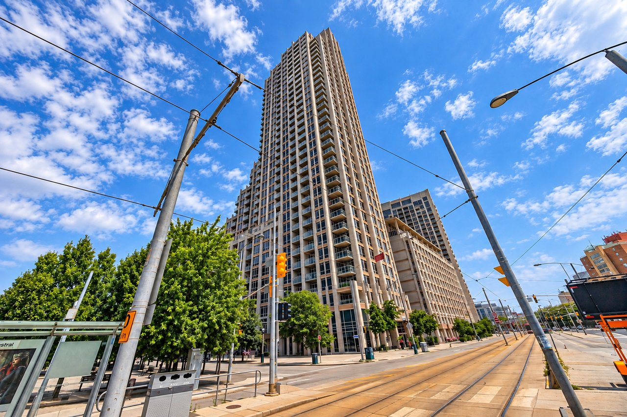 Tall modern high-rise with balconies beside an urban street with tram tracks and overhead wires.