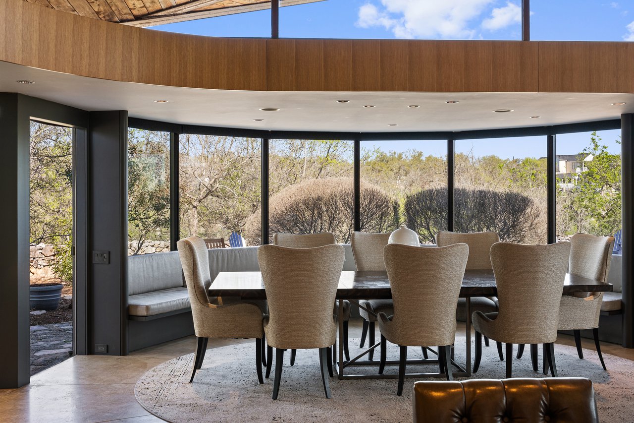 Formal dining room wrapped in a curved glass wall with built-in window bench seating and hill country views, 4002 Tyx Trail, Spicewood, Texas.